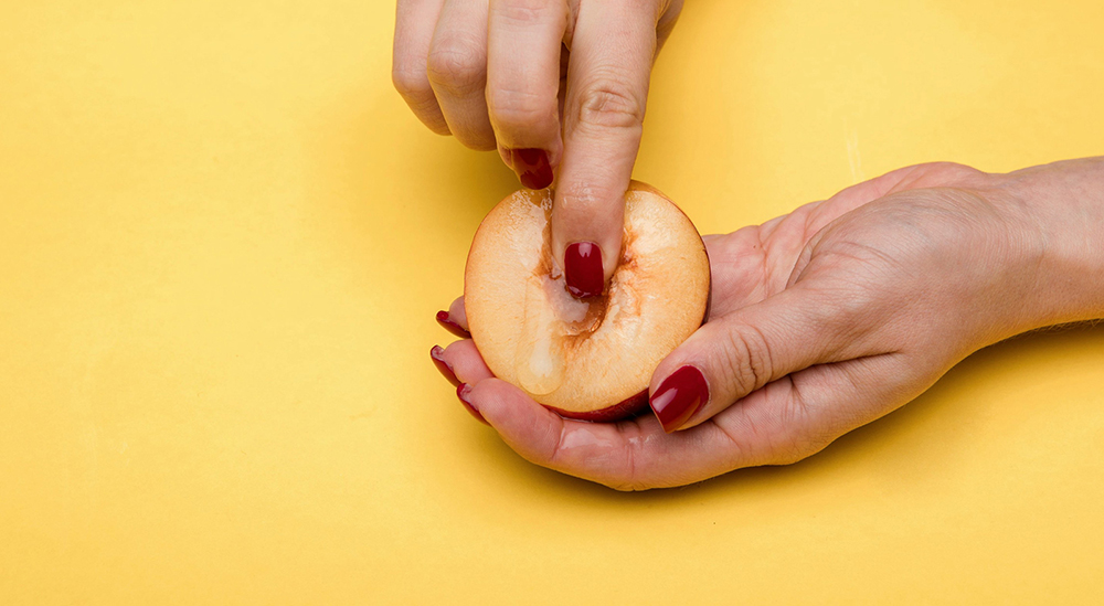 노란색 배경에서 한 손으로는 자두를 들고, 한 손으로는 자두의 중앙을 만지고 있다