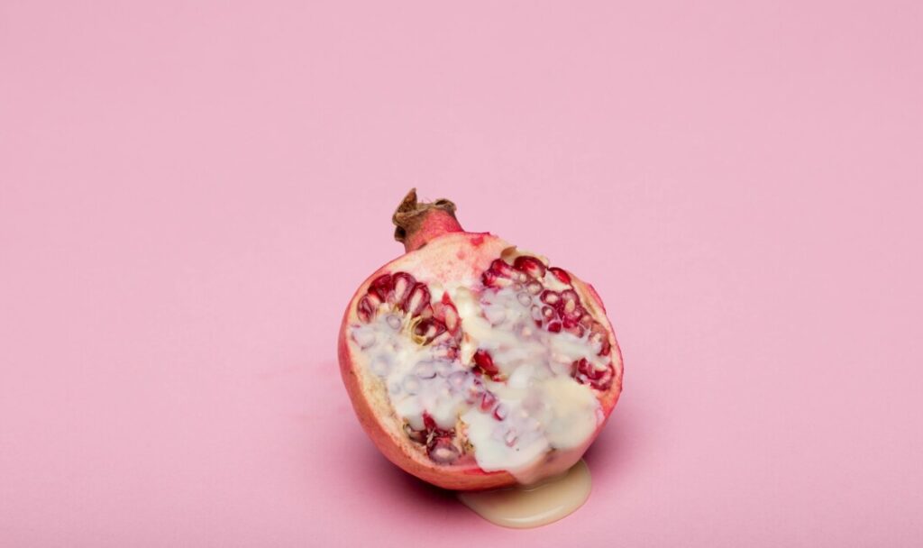 분홍색 배경 위에 반으로 잘린 석류가 놓여 있고, 그 위로 하얗고 점성이 있는 액체가 흘러내려 바닥에 고여 있다.